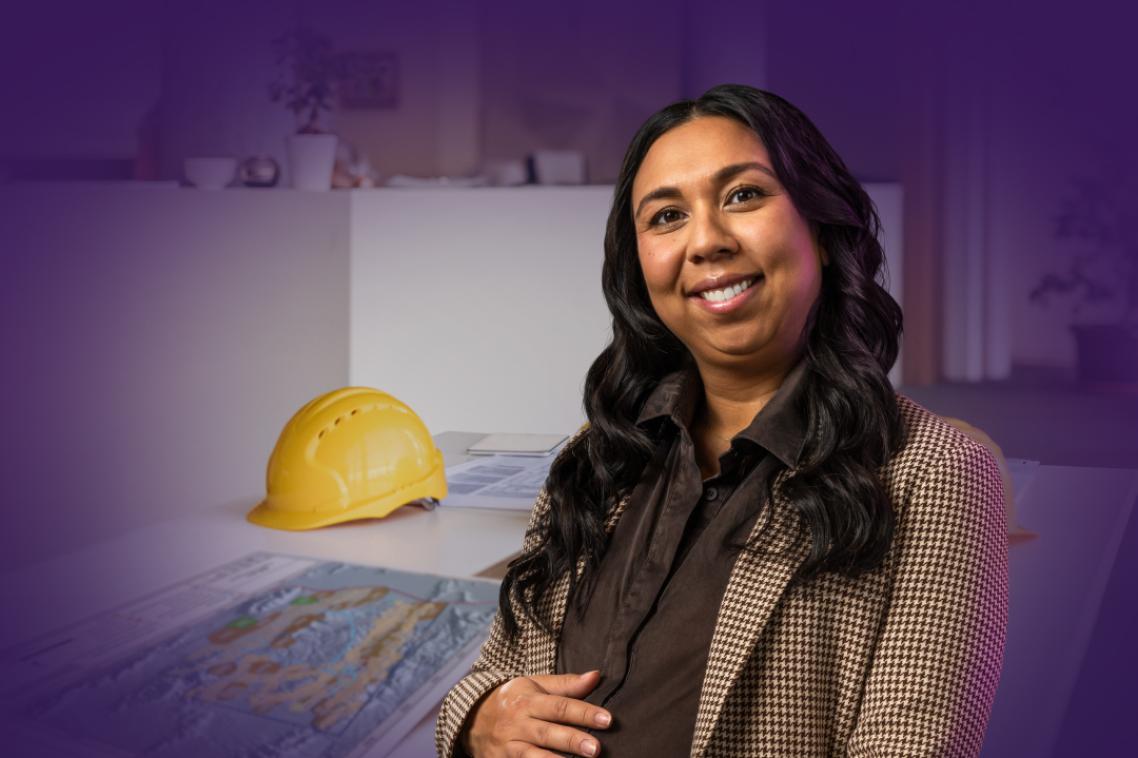 Women smiling in jacket stands against desk with a plan and yellow hard hat on top.